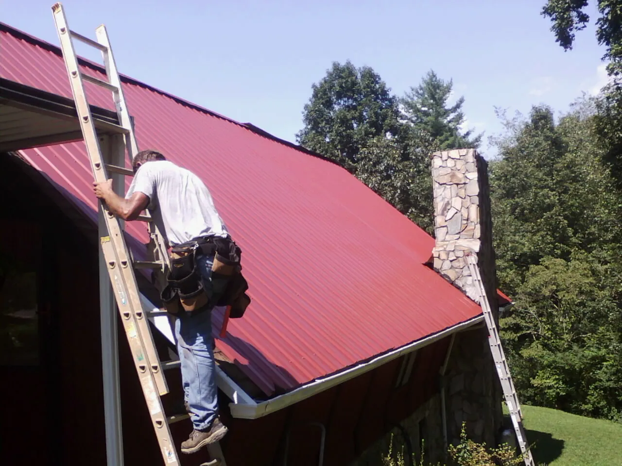 Roofer on a ladder beside a red metal roof and stone chimney — Soffit & Fascia Repair by Roofing Brothers in Powhattan